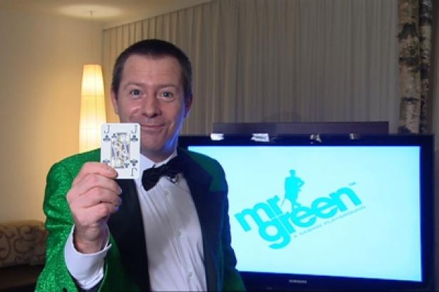 Mr Green-Show mit Alex Krause/Mr Green Österreich Ein Mann im grünen Jackett präsentiert eine Spielkarte vor einem Bildschirm mit dem Mr. Green Logo.