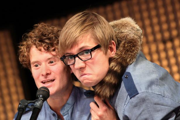 Zwei Männer stehen zusammen, einer mit lockigem Haar und Mikrofon, der andere mit Brille und Fellkragen.