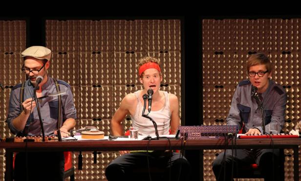 Drei Männer sitzen an einem Tisch mit Musikinstrumenten und Mikrofonen auf einer Bühne.