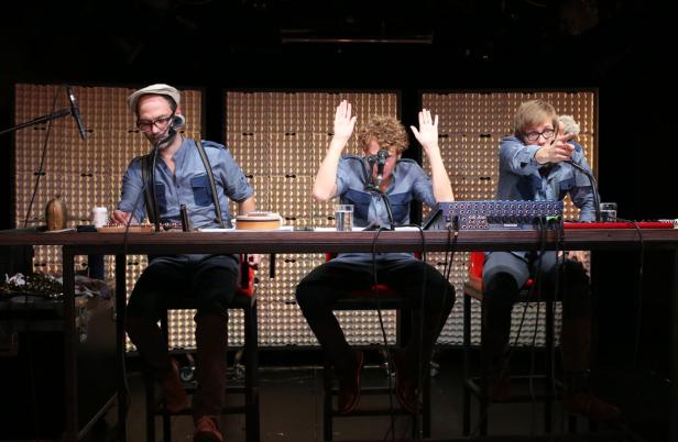 Drei Männer sitzen an einem Tisch mit Mikrofonen und Musikinstrumenten vor einem goldenen Hintergrund.