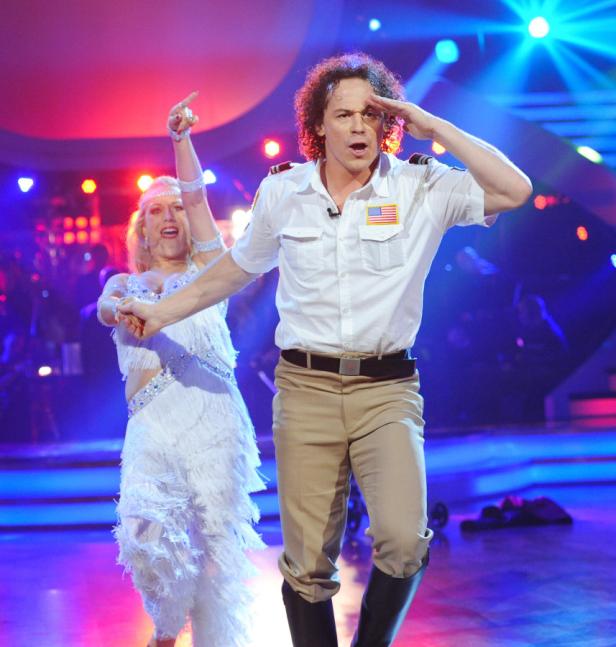 Ein Mann in Uniform und eine Frau im weißen Kleid tanzen auf einer Bühne.