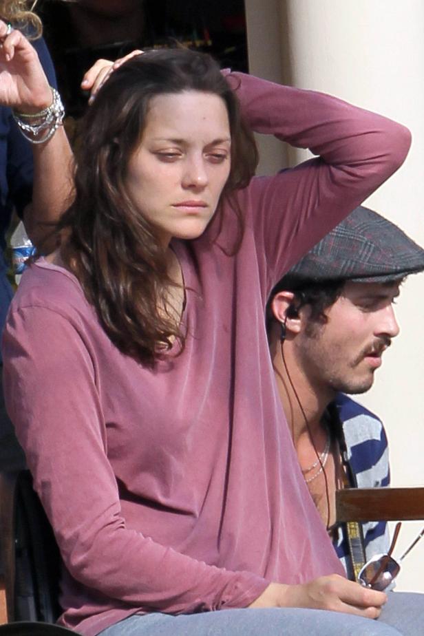 Marion Cotillard richtet ihre Haare, während ein Mann mit Schirmmütze im Hintergrund sitzt.