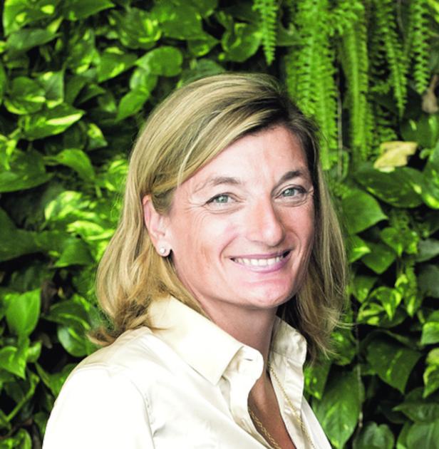 Eine Frau mit blonden Haaren lächelt vor einem grünen Blätterhintergrund.