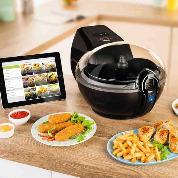Eine Heißluftfritteuse der Marke Tefal steht neben einem Tablet und zwei Tellern mit frittierten Speisen.