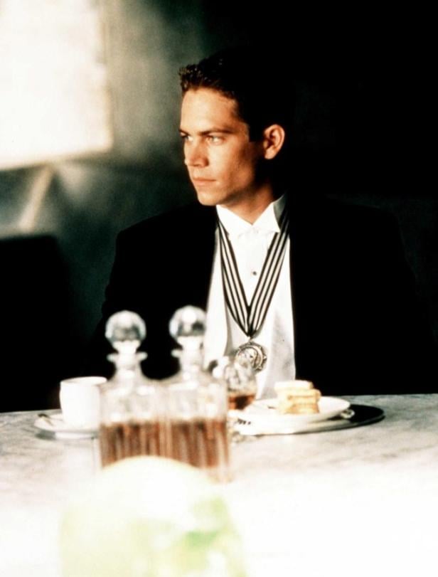 Paul Walker in Anzug mit Orden sitzt an einem Tisch mit Getränken und Speisen.