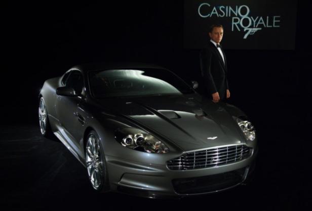 Daniel Craig posiert neben einem Aston Martin DBS vor einem „Casino Royale“-Plakat.