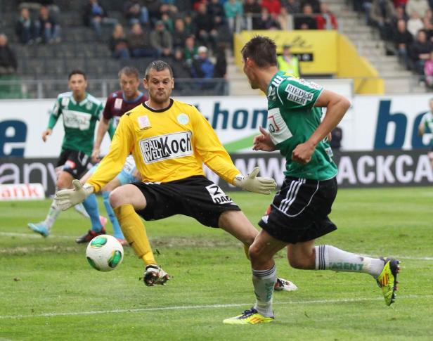 Ein Fußballtorwart wehrt einen Schuss ab, während ein anderer Spieler an ihm vorbeiläuft.