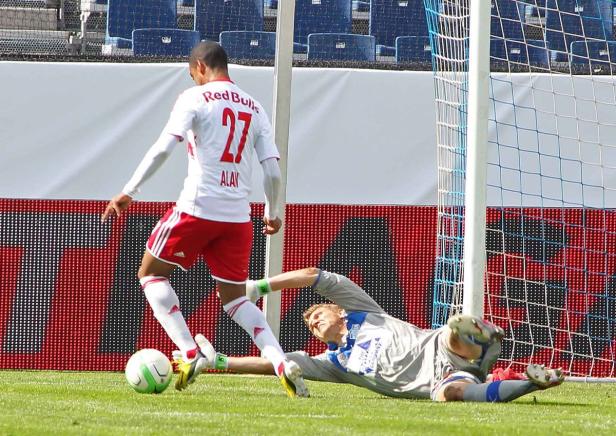 Ein Fußballspieler in Rot schießt auf das Tor, während der Torwart sich streckt, um den Ball abzuwehren.