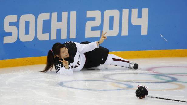 Eine Eishockeyspielerin liegt auf dem Eis vor dem Schriftzug „Sochi 2014“ und zeigt ein Victory-Zeichen.