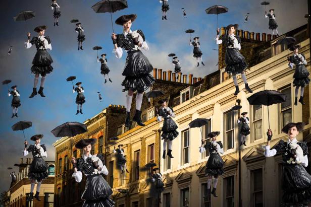 Eine Frau im extravaganten Kleid schwebt mit einem Regenschirm vor einer Häuserfassade.