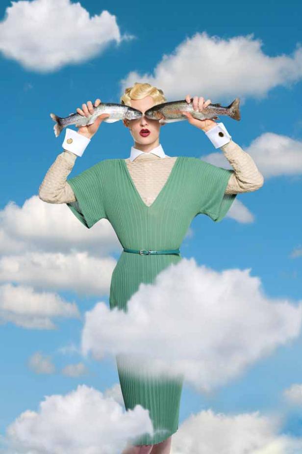 Eine Frau hält zwei Fische vor ihre Augen, vor einem Himmel mit Wolken.