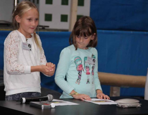 Zwei junge Mädchen stehen an einem Tisch mit Mikrofon und Papieren.