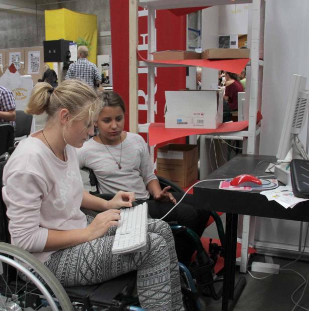 Zwei junge Frauen sitzen in Rollstühlen und arbeiten an einem Computer.