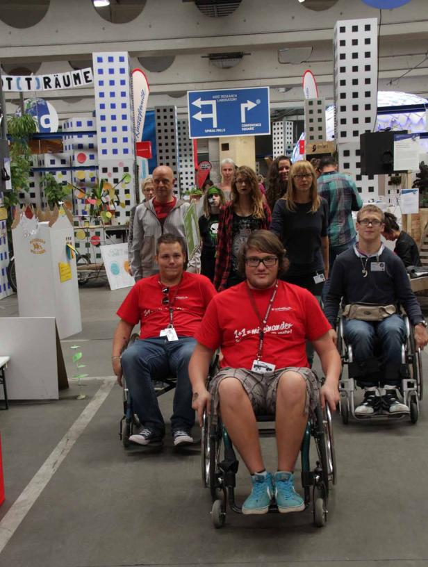 Eine Gruppe von Menschen, darunter drei im Rollstuhl, posiert vor einem farbenfrohen Hintergrund mit Schildern.