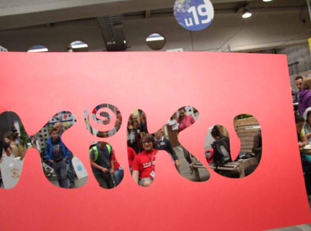 Eine rote Pappwand mit dem ausgeschnittenen Wort „kiku“, durch das Menschen zu sehen sind.