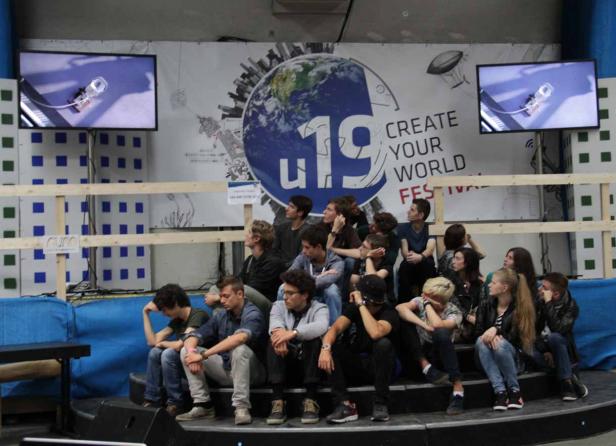 Eine Gruppe junger Leute sitzt vor einer Bühne mit Bildschirmen und dem Schriftzug „Create Your World Festival“.