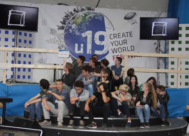 Eine Gruppe junger Leute sitzt auf einer Bühne vor einem Banner mit der Aufschrift „Create Your World Festival“.