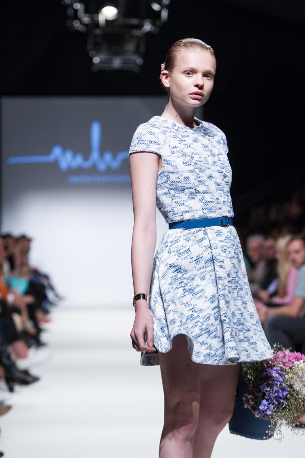 Ein Model präsentiert ein blau-weißes Kleid mit blauem Gürtel auf einem Laufsteg.