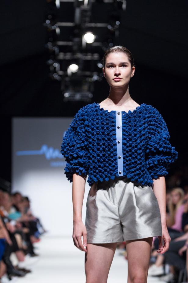 Eine Frau präsentiert ein blaues Oberteil mit Blumenmuster und silberne Shorts auf einem Laufsteg.