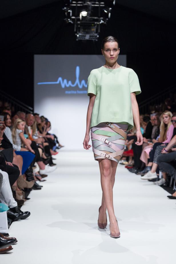 Eine Frau präsentiert ein grünes Oberteil und einen Rock mit Gürtel auf einem Laufsteg.