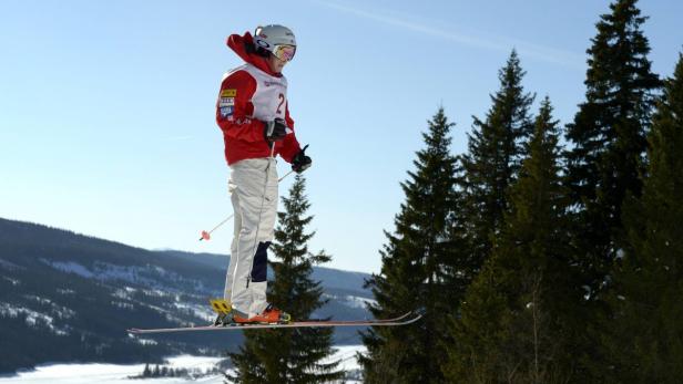 Ein Skispringer in roter Jacke und weißer Hose springt vor einer Waldkulisse.