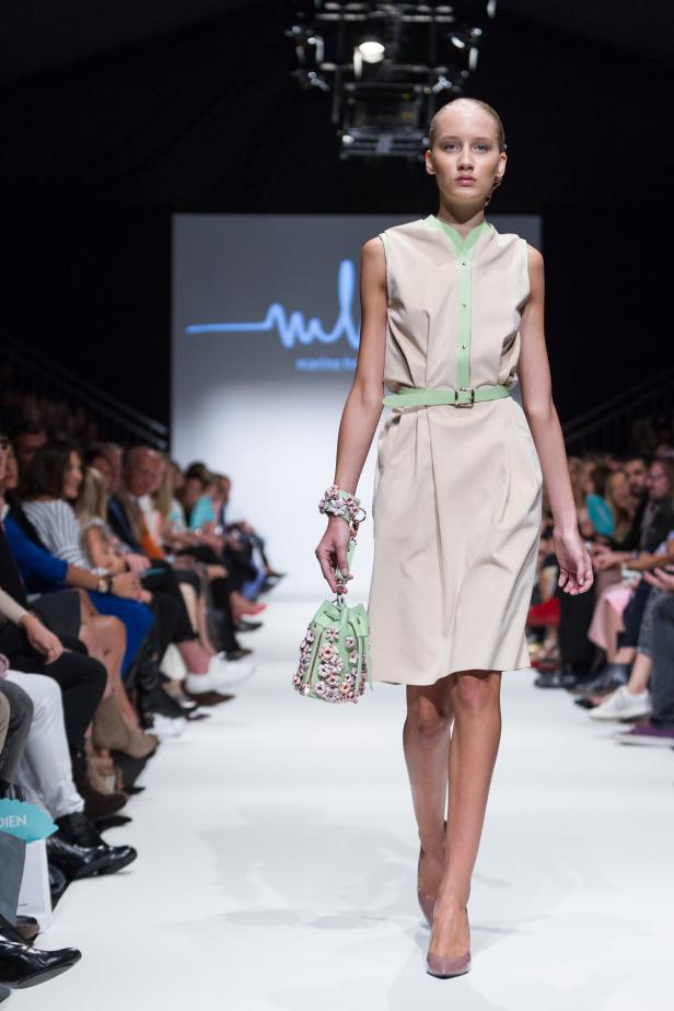 Eine Frau präsentiert ein beiges Kleid mit hellgrünem Gürtel und eine passende Handtasche auf dem Laufsteg.