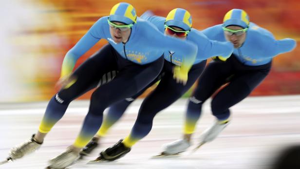 Drei Eisschnellläufer in blauen Anzügen und gelben Mützen auf einer Eisbahn.