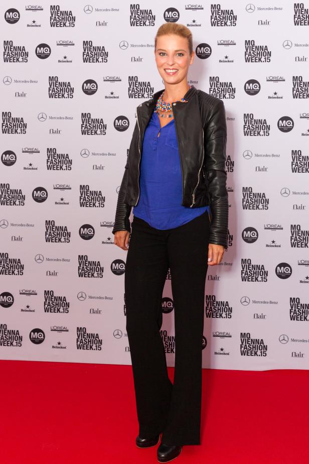 Eine Frau in Lederjacke posiert vor einer Wand mit Logos der Vienna Fashion Week.