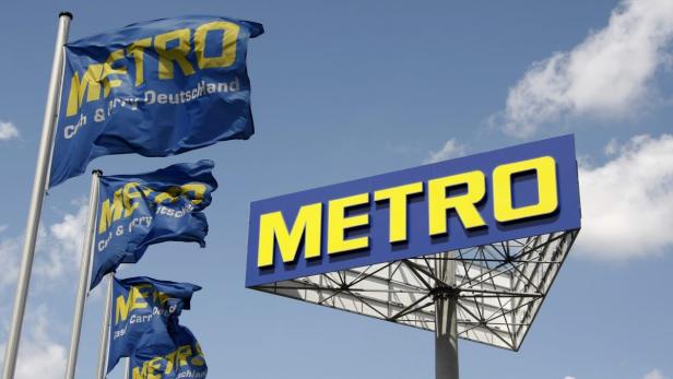 Ein Schild und mehrere Flaggen mit dem Logo von Metro Cash & Carry vor blauem Himmel.