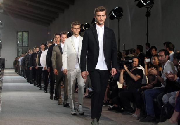 Eine Reihe von männlichen Models präsentieren Herrenmode auf einem Laufsteg.