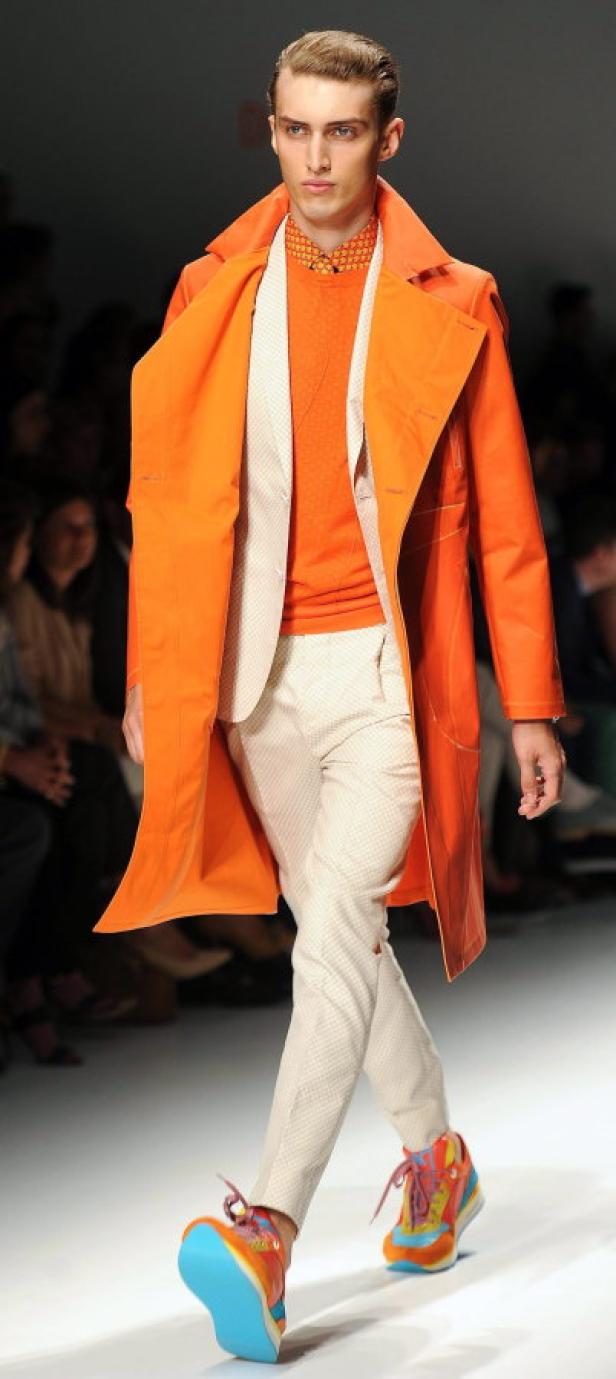 Ein Mann in einem orangefarbenen Mantel und bunten Turnschuhen läuft über einen Laufsteg.