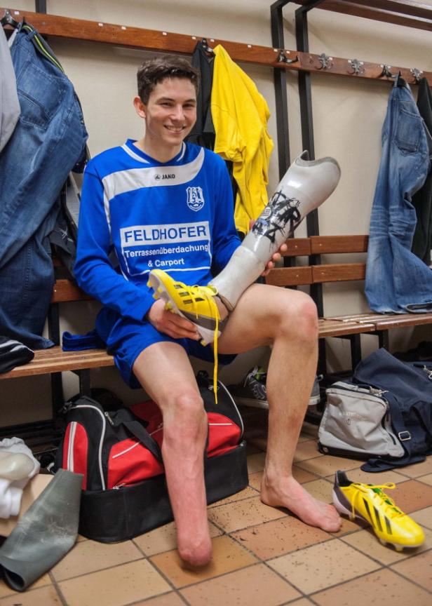 Ein lächelnder Fußballspieler mit Beinprothese sitzt in der Umkleide und hält einen Fußballschuh.