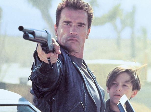Arnold Schwarzenegger zielt mit einer Schrotflinte, neben ihm steht ein Junge.