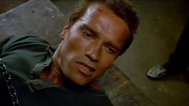 Arnold Schwarzenegger liegt gefesselt und verletzt auf einer Werkbank.