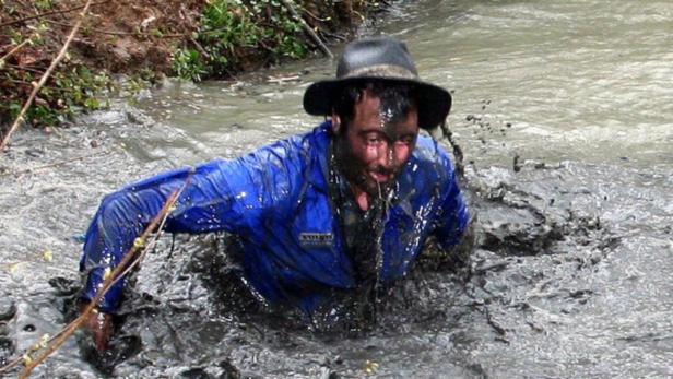 Ein Mann mit Hut und blauer Jacke watet durch schlammiges Wasser.