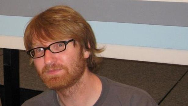 Ein Mann mit rötlichem Haar und Bart trägt eine schwarze Brille.