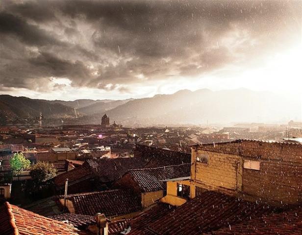 Blick über eine Stadt mit roten Ziegeldächern im Regen, im Hintergrund Berge.
