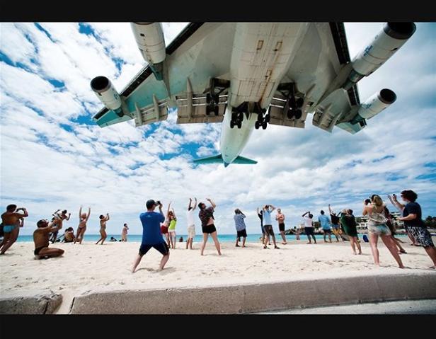Ein großes Flugzeug landet über einem Strand, wo viele Menschen es beobachten und fotografieren.