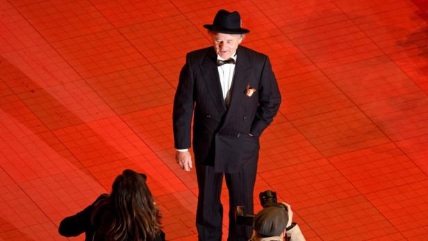 Ein älterer Mann mit Hut und Anzug posiert auf einem roten Teppich für Fotografen.