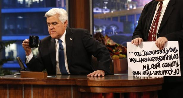 Jay Leno sitzt an einem Schreibtisch und hält eine Tasse in der Hand, während jemand ein Schild mit einer Nachricht hält.