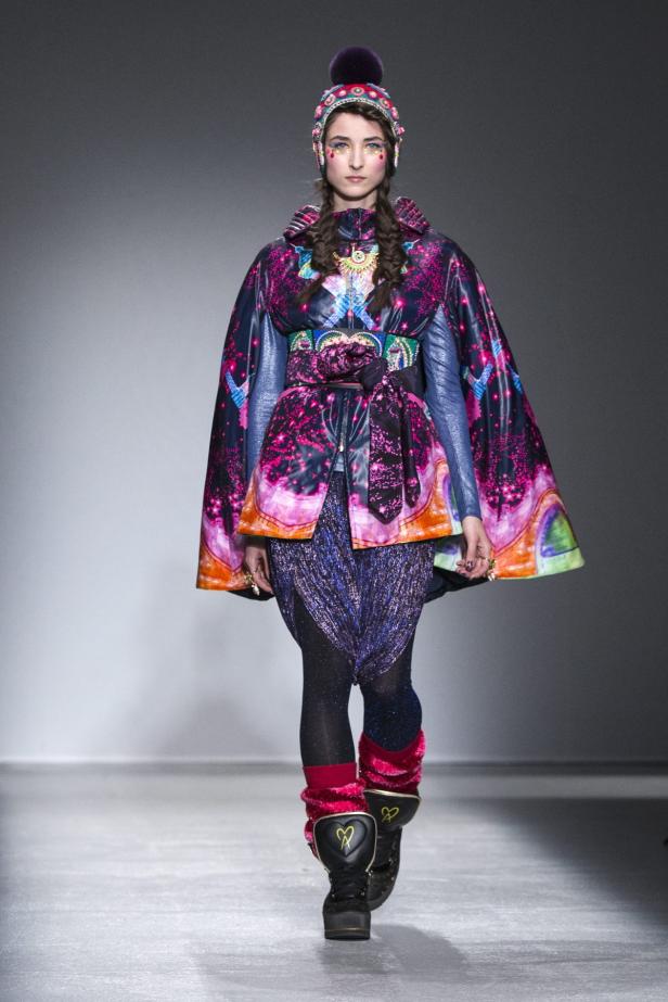 Ein Model präsentiert ein farbenfrohes Outfit mit Cape, Mütze und Plateaustiefeln auf dem Laufsteg.