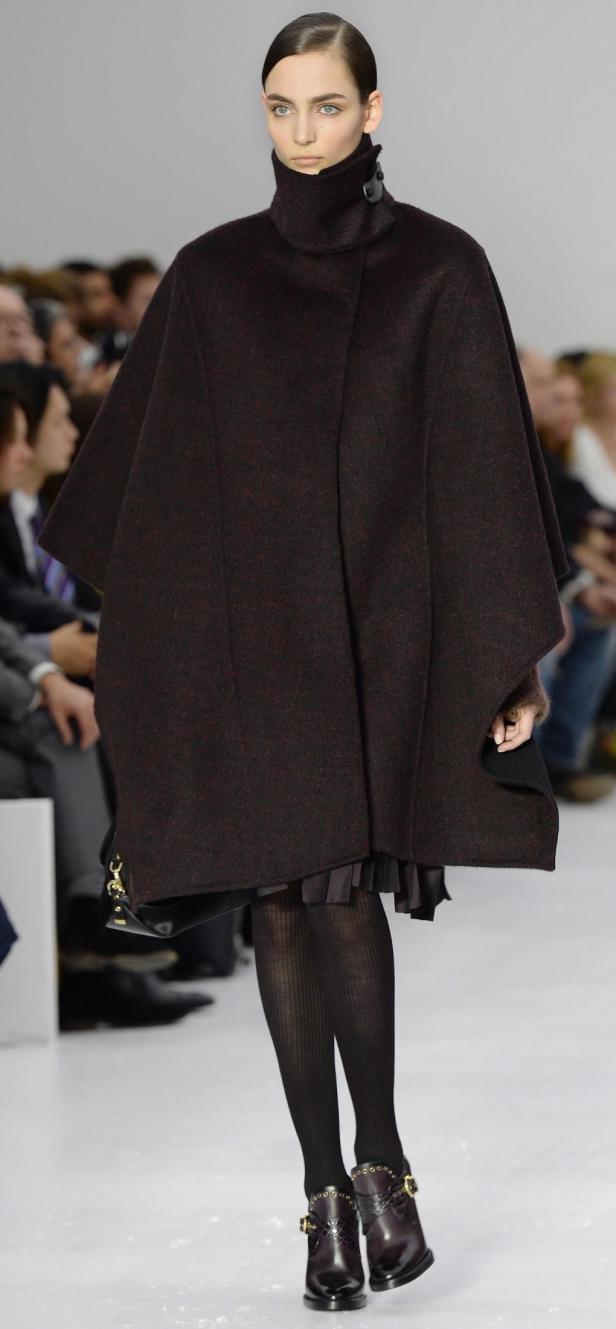 Eine Frau auf dem Laufsteg trägt einen braunen Cape-Mantel, Strumpfhosen und Stiefeletten.