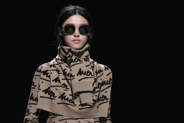 Eine Frau mit Sonnenbrille trägt ein beiges Strickoutfit mit schwarzer Aufschrift.