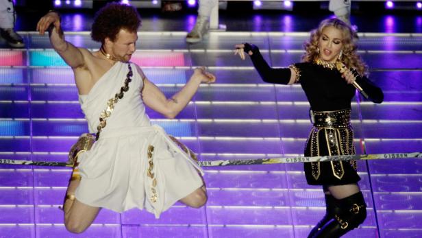 Madonna tritt mit einem Tänzer in einem weißen, griechisch inspirierten Kostüm auf einer Bühne auf.