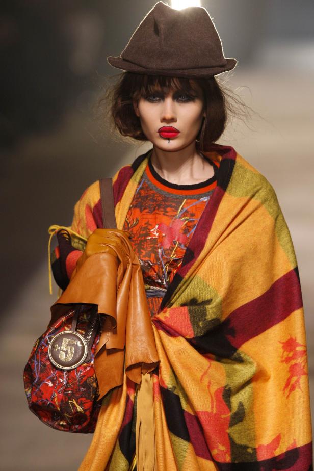 Eine Frau mit Hut und auffälligem Make-up präsentiert ein farbenfrohes Outfit mit Tasche.