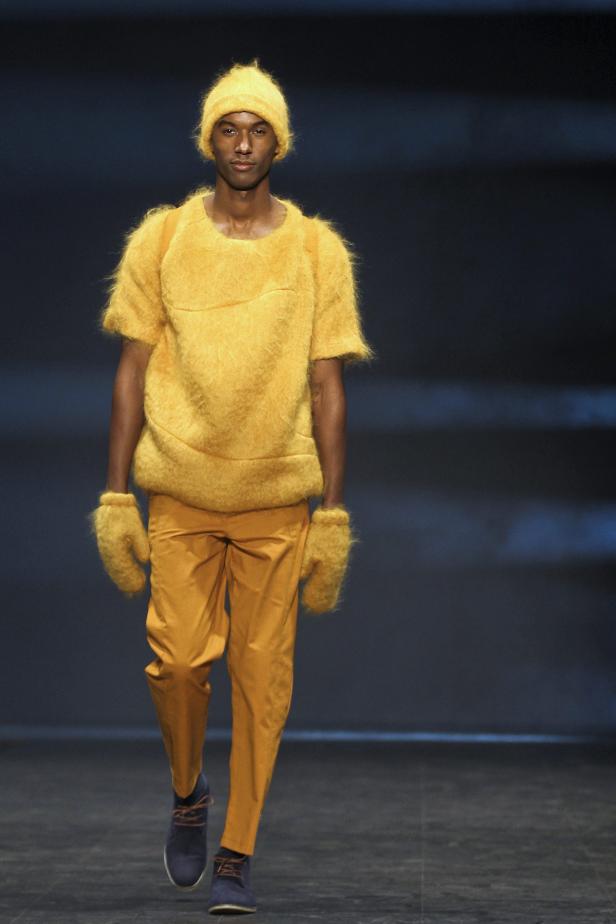 Ein Mann in gelber Kleidung und Mütze auf einem Laufsteg.