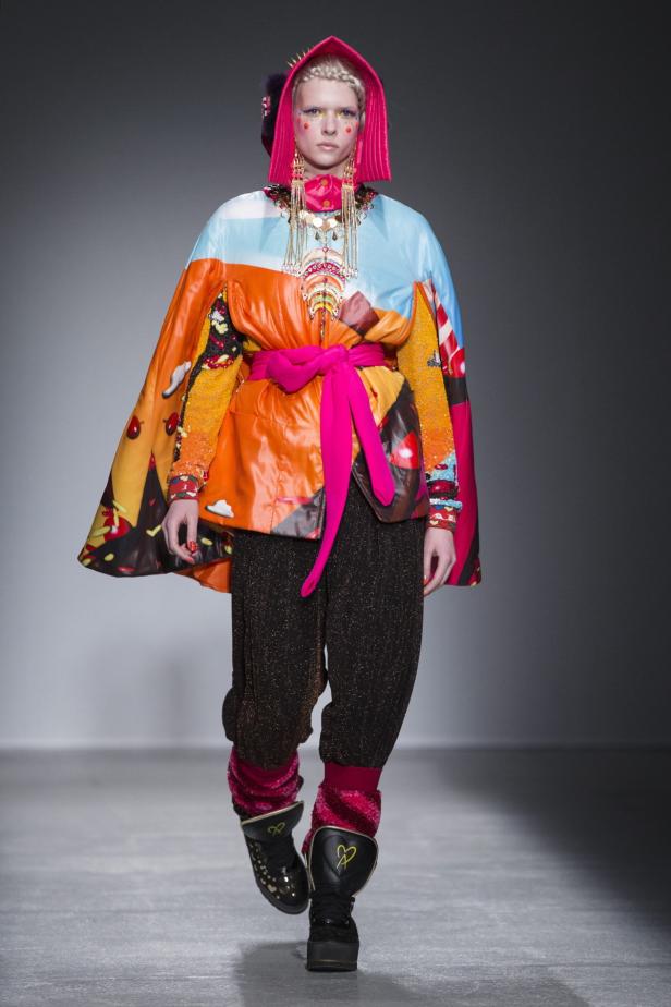 Ein Model präsentiert ein farbenfrohes Outfit mit Cape und auffälligem Schmuck auf dem Laufsteg.
