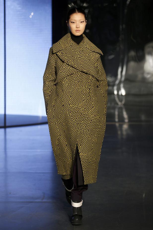 Eine Frau mit strengem Dutt trägt einen gemusterten Mantel auf dem Laufsteg.