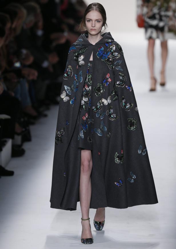 Ein Model trägt einen grauen Umhang mit bunten Schmetterlingsapplikationen auf dem Laufsteg.