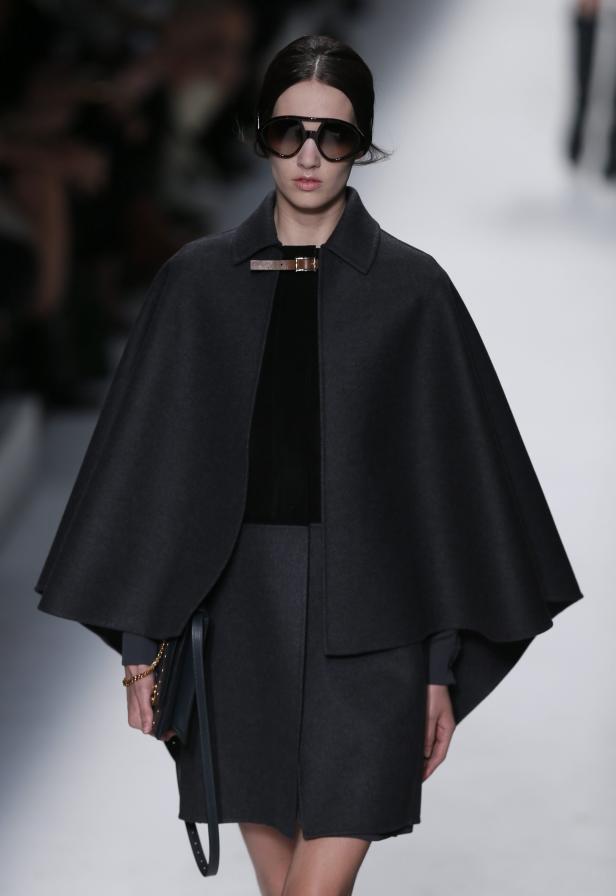 Eine Frau mit Sonnenbrille trägt einen grauen Cape-Mantel und eine Handtasche.